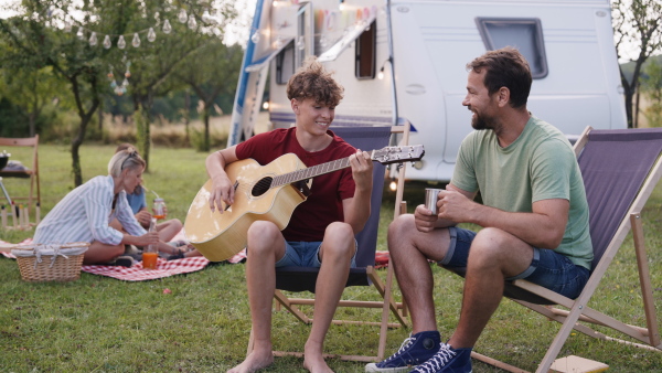 Family traveling in a caravan, camping and exploring new places. Father teaching boy to play guitar, sitting on folding chairs.
