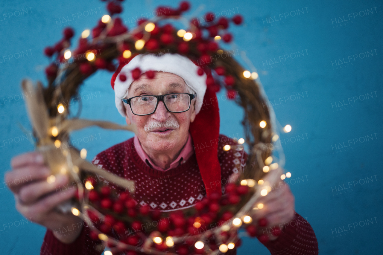 Portrait of happy senior man with Santa Claus hats in studio shoot, looking trough Christmas wreath.