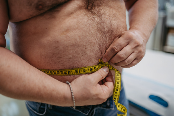 Close up of overweight man measuring waist, using tape measure. Obesity affecting middle-aged men's health. Concept of health risks of overwight and obesity.