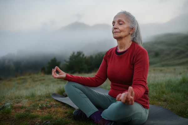 A senior woman doing breathing exercise in nature on early morning with fog and mountains in background.