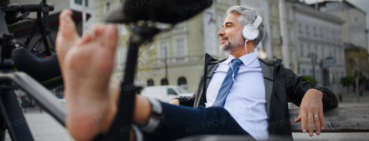 A businessman with bike sitting on bench, listening to music with feet up and resting. Commuting and alternative transport concept