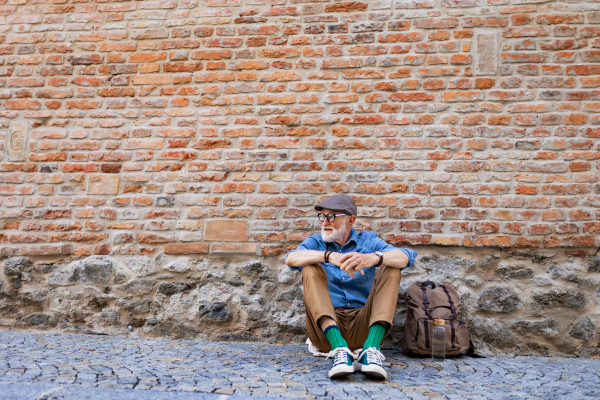 Portrait of senior tourist exploring new city, interesting places. Elderly man with backpack sitting by brick wall, resting. Traveling and solo trips in retirement, copy space.