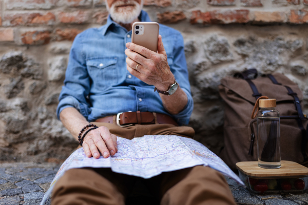 Senior tourist exploring a new city, interesting places. Elderly man holding a paper map and smartphone, looking for the route. Traveling and trips in retirement.