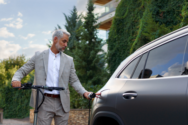 Businessman in suit on way to work standing at an eletric scooter and charging his electric car. Concept of eco commuting and green transportation.