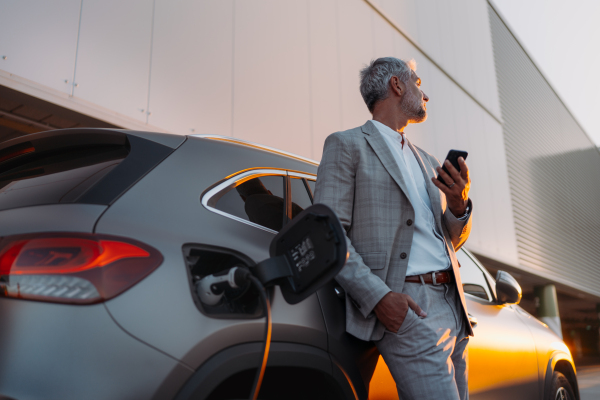A businessman holding smartphone while charging car at electric vehicle charging station, closeup.