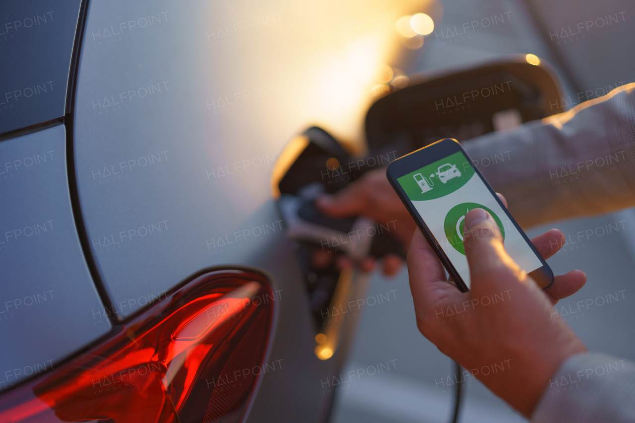 A man holding smartphone while charging car at electric vehicle charging station, closeup.