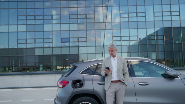 Mature businessman in suit charging battery of electric car while using mobile phone in front of modern office building.
