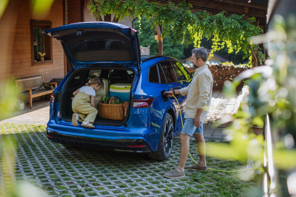 Father charging their electric car, little daughter sitting and waiting in car trunk.