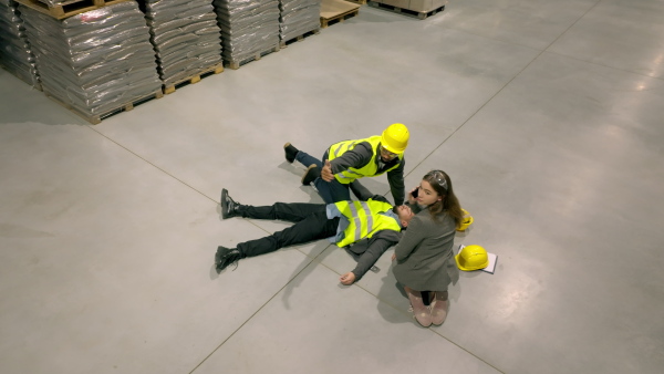 Top view of colleagues giving first aid to unconscious worker after accident at work in warehouse, modern factory. Calling the ambulance. Concept of occupational safety and health in workplace.