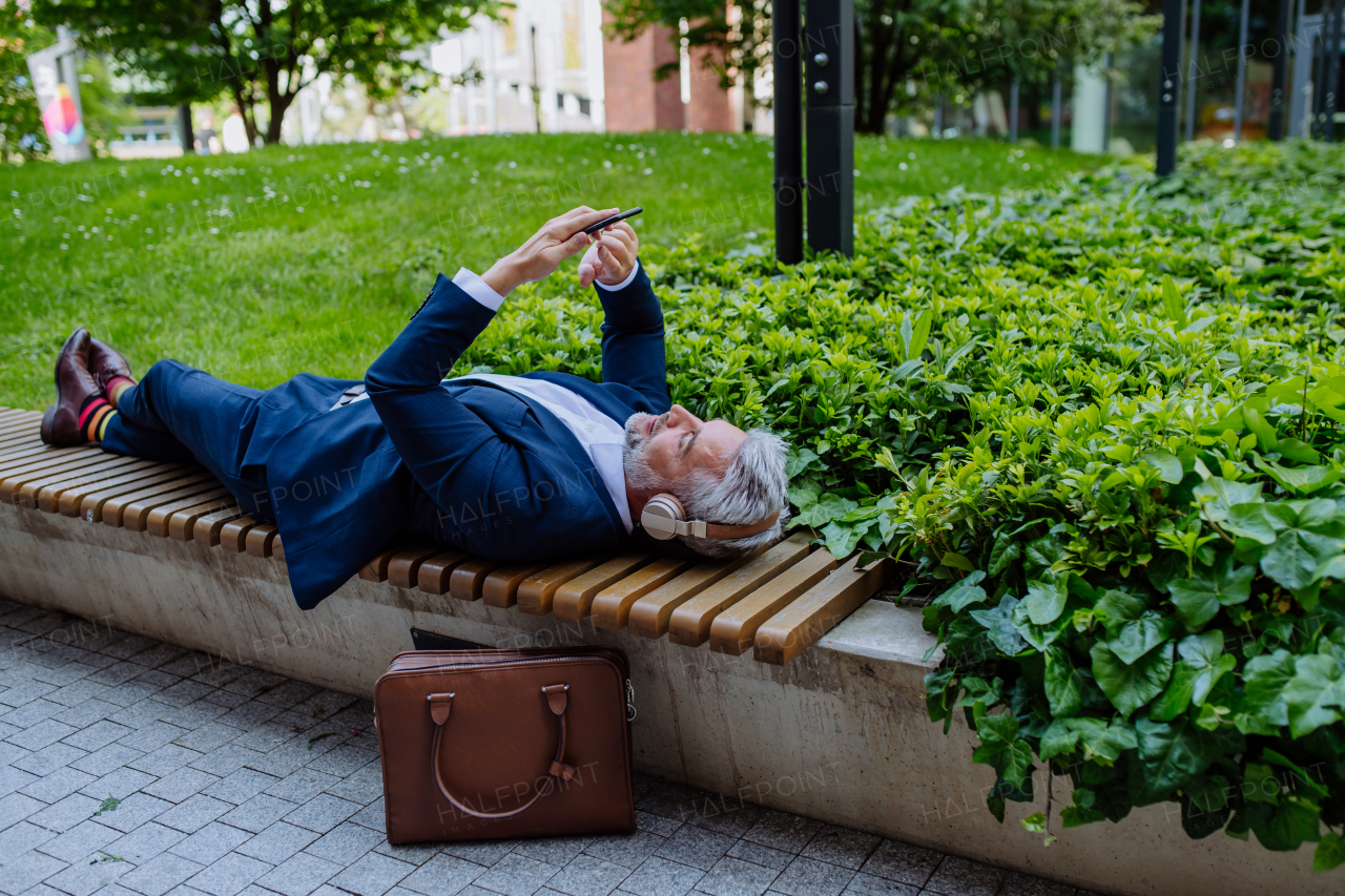 A mature businessman enjoying music in headphones relaxing on bench in city park during break at work.