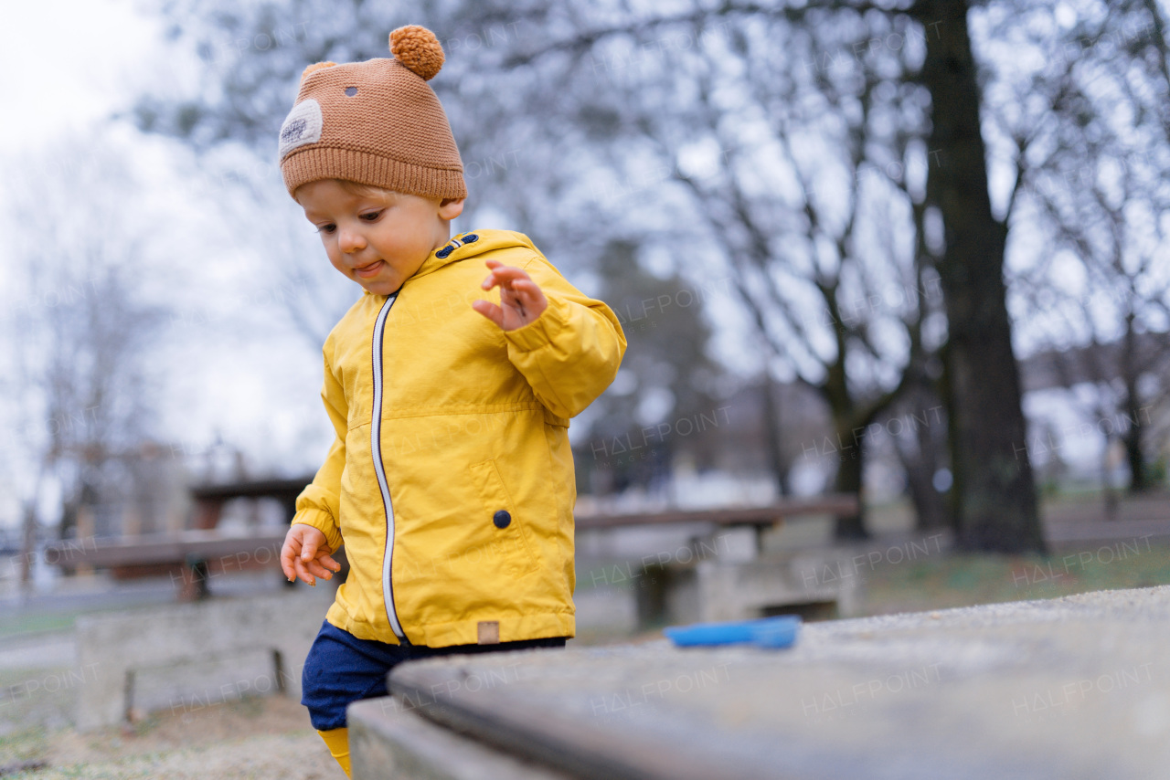 Happy little boy with a teddy bear cap playing on playground at sandpit during autumn day.