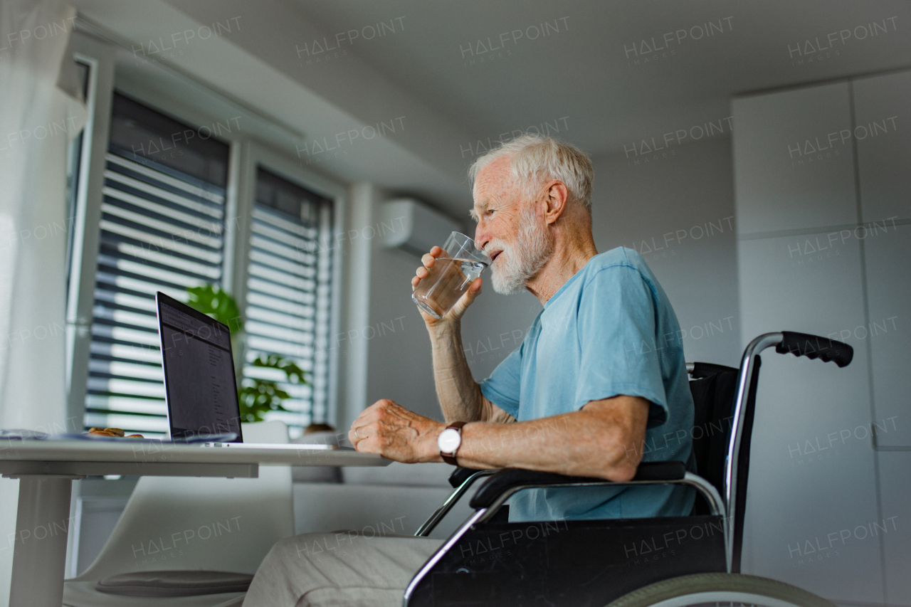Senior man in a wheelchair shopping online on laptop. Elderly man using digital technologies, working on a notebook in his kitchen. Concept of seniors and digital skills.