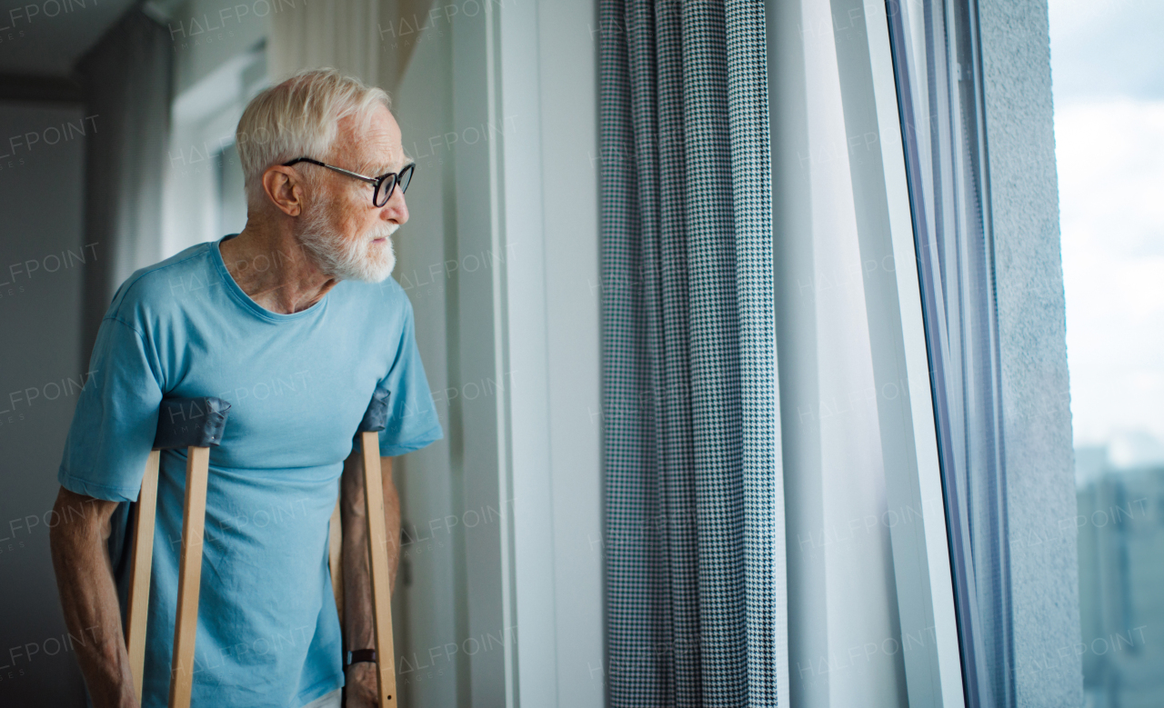 Sad senior man with crutches spending time alone in his apartment, looking out of window. Concept of loneliness and dependence of retired people.