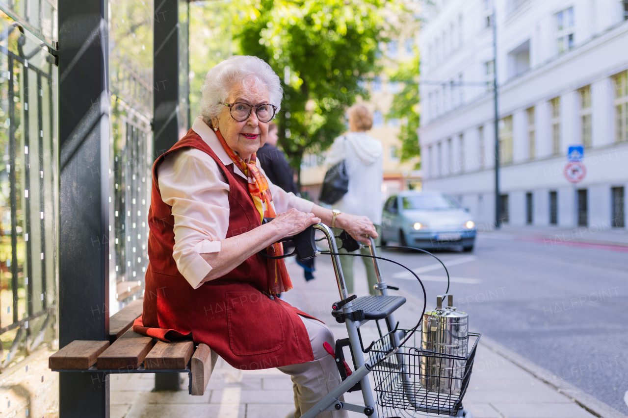 Senior woman in city, sitting on a bus stop, waiting for public transport, rollator next to her.