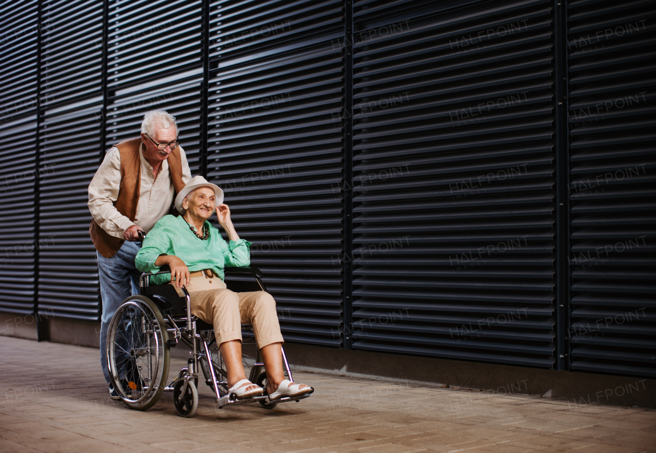 Portrait of senior couple with woman in wheelchair. Elderly man pushing his lovely wife in wheelchair. Concept of chronic illness in elderly couple. Portrait with copy space on black background. Senior siblings on a Sunday stroll in the city.