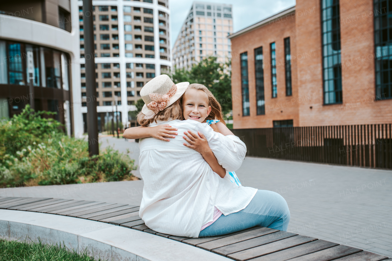 Grandma sitting on bench with granddaugter in the city, embracing her. Girl spending time with senior grandmother, during summer break or after school.
