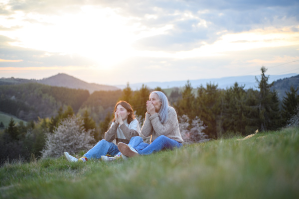 A happy senior grandmother with teenage granddaguhter sitting and whistling on grass in nature on spring day.