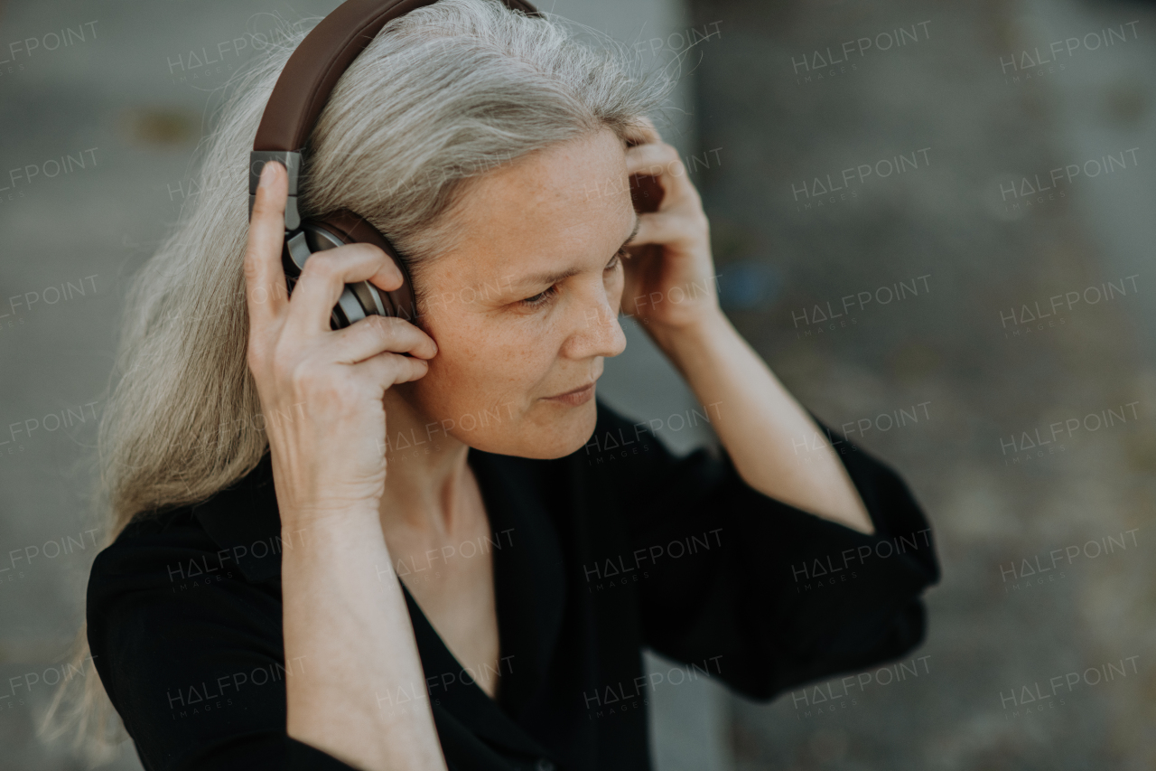 Head shot of a beautiful woman with gray hair, sitting on concrete stairs in the city, listening to music through wireless headphones.