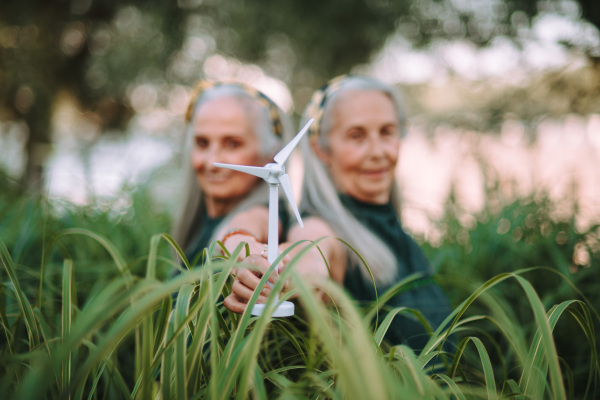 Senior women, twins holding a plastic model of wind turbine outdoor in nature, concept of future, ecology and renewable resources, message for next generation.