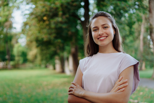 Portrait of a beautiful young woman standing outside in a city park, with copy space. The smiling woman standing with her arms crossed, looking at camera.