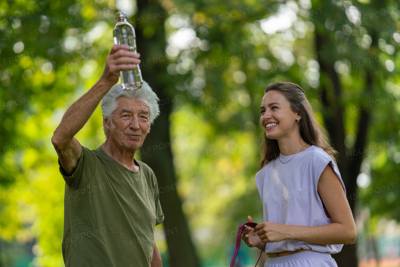 The senior man drinking water during workout, staying hydrated. Young physiotherapist training outdoors in the park with senior patient, letting him rest after exercising.