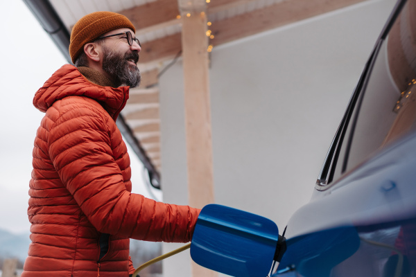 Man charging electric car during cold snowy day. Hansome mature man putting charger in charging port during cold weather. Charging and driving electric vehicles during winter season. Low angle shot.