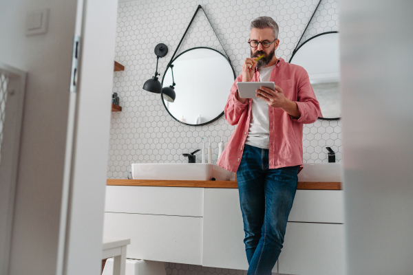 Man shopping online while brushing his teeth in bathroom. Holding tablet, scrolling and buying in eshop with toothbrush in mouth.