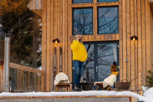 Mature man phone calling from patio, drinking hot tea, coffee and enjoying beautiful winter day. Handsome man spending relaxing, stress-free winter weekend in cabin in mountains, enjoying alone time.