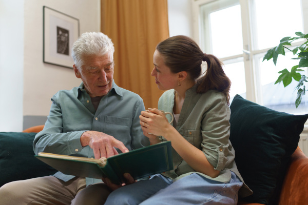 Grandfather with his granddaughter looking at family photo album. Nostalgic senior man showing photos of himself from his youth, remebering old days through pictures.