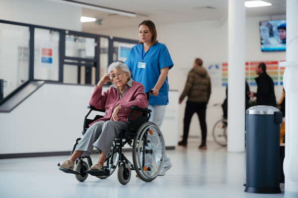 Portrait of nurse pushing senior patient in a wheelchair across hospital corridor, hall. Emotional support for elderly woman.