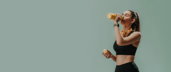 A young sporty woman in sportswear with wireless headset drinking from bottle over green background, copy space