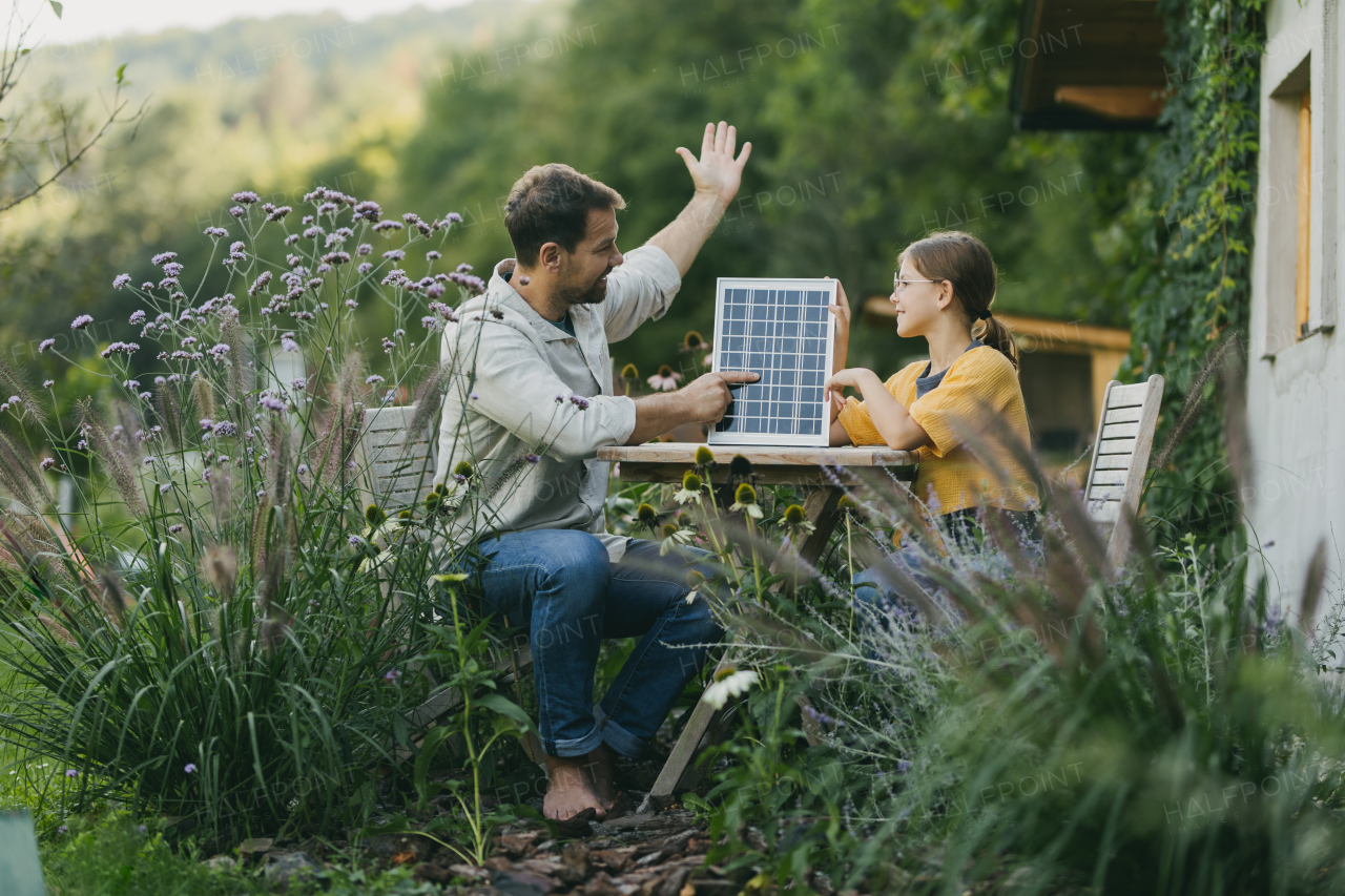 Father with daughter sitting outdoors in the garden, with model of solar panel. Solar energy and sustainable lifestyle of young family. Concept of green energy and sustainable future for next generations.