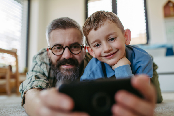 Little boy making selfie on smartphone with father, lying on the floor in kids room, making silly faces. Dad explaining technology to son, digital literacy for kids.