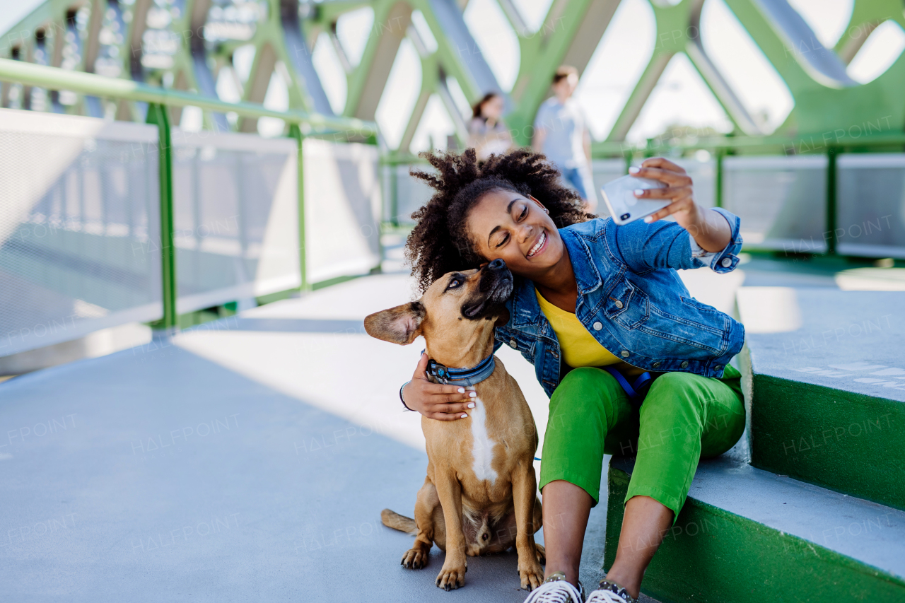 Multiracial girl sitting and resting with her dog outside in the bridge, taking selfie with him, spending leisure time together. Concept of everyday life with pet.