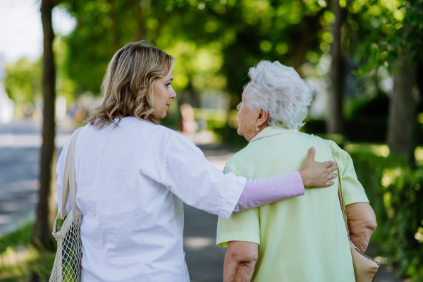 A rear view of caregiver with senior woman on walk with walker in park with shopping bag.