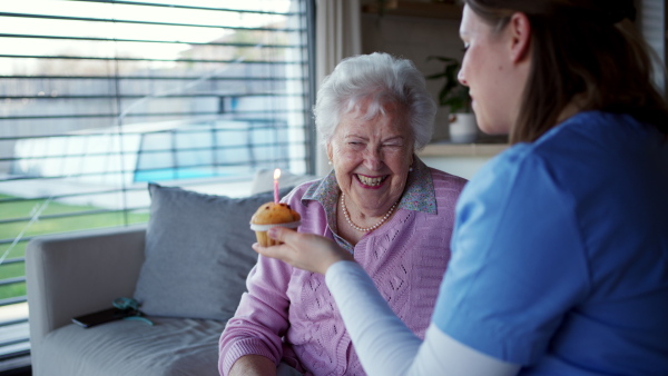 Elderly celebrating birthday with home caregiver, blowing candle on sweet muffin cake. Senior woman at home is thankful for care and assistance from nurse.