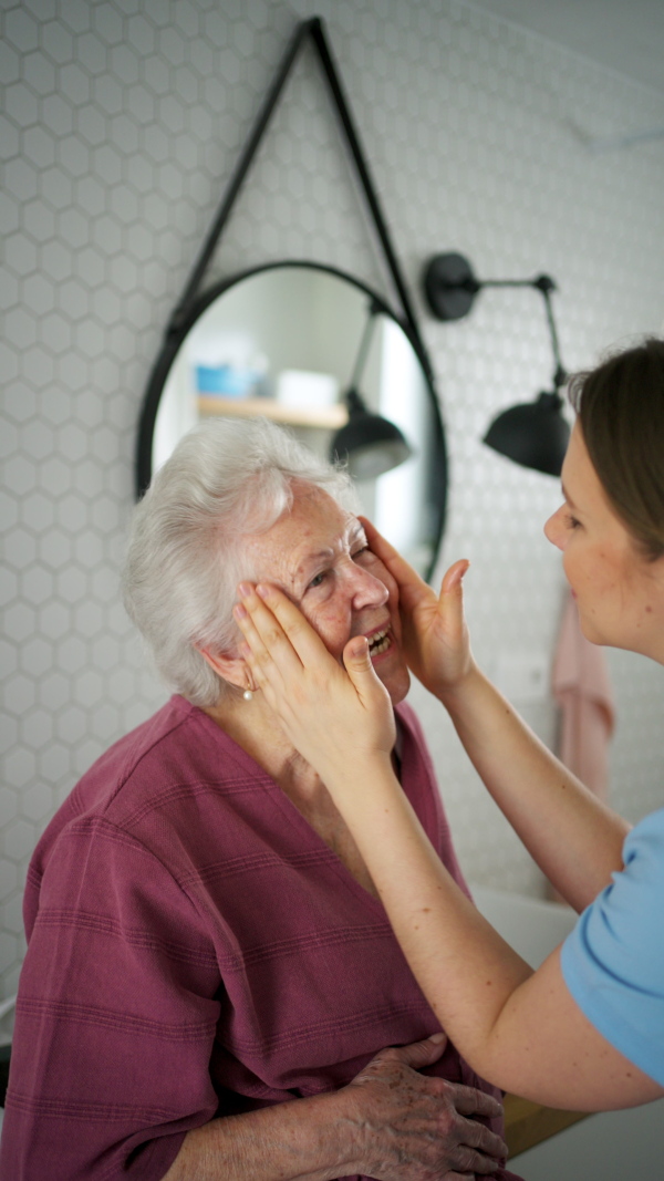 Home caregiver helping elderly woman with skincare in bathroom, applying moisturise cream on face. Senior woman at home with young beautiful nurse.