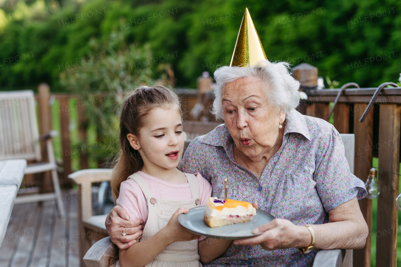 Granddaughter celebrating birthday with elderly grandma, blowing candle on cake. Weekend in cottage.
