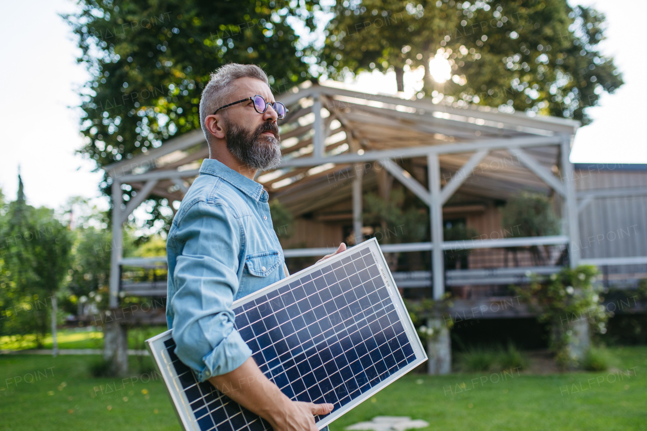 Man looking at solar panel in arms, standing in garden. Sustainable lifestyle and green household, home.