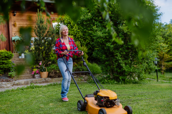 An ederly woman mowing grass with lawn mower in the garden, garden work concept.