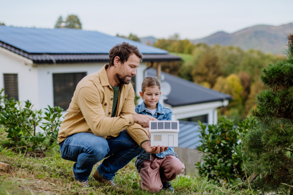 Little girl with her father holding paper model of house with the solar panels, explaining how it works.Alternative energy, saving resources and sustainable lifestyle concept.