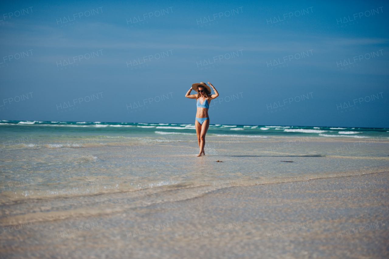 Portrait of a beautiful slim woman standing on the beach in a bikini and a straw hat. Full body shot of woman in blue swimsuit, sunglasses enjoying sandy beach and crystalline sea of Mnemba beach in Zanzibar, with copy space. Concept of beach summer vacation.