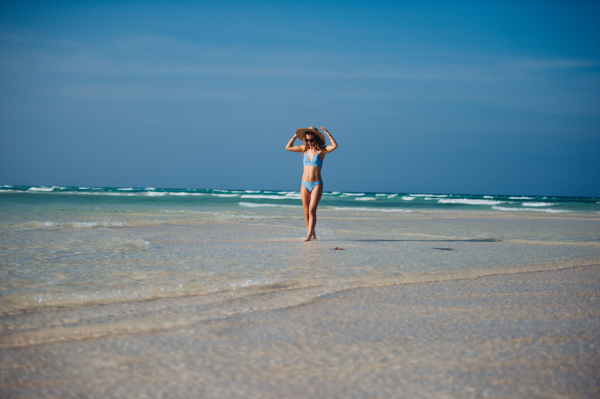 Portrait of a beautiful slim woman standing on the beach in a bikini and a straw hat. Full body shot of woman in blue swimsuit, sunglasses enjoying sandy beach and crystalline sea of Mnemba beach in Zanzibar, with copy space. Concept of beach summer vacation.