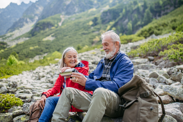 Active elderly couple hiking together in autumn mountains, on senior friendly trail. Husband and wife rehydrating, enjoying warm tea, coffee from thermos. Senior tourist with backpack getting rest during hike.