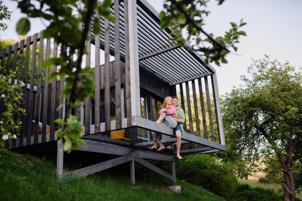 A young couple sitting and cuddling in hammock terrace in their new home in tiny house in woods, sustainable living concept.