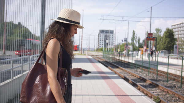Young woman commuter waiting for public transport, looking at application on her mobile phone.
