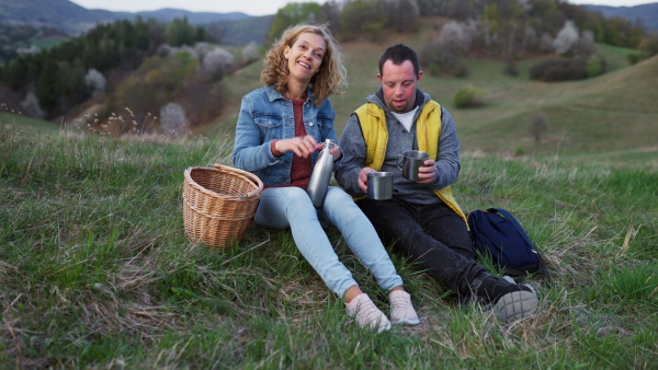 Happy young man with Down syndrome with his mother resting in nature, sitting and drinking tea from a thermos.