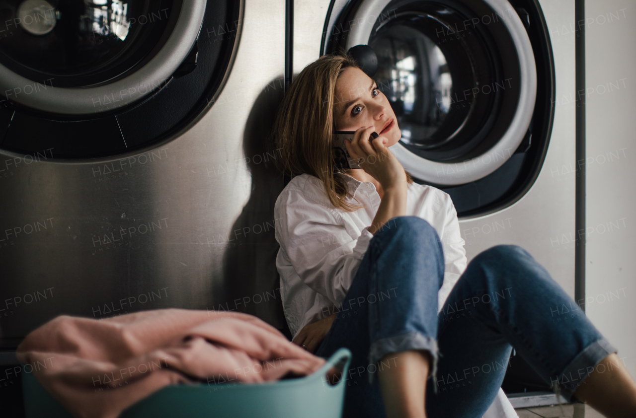 Young woman calling with smartphone, sitting in a laundry room.