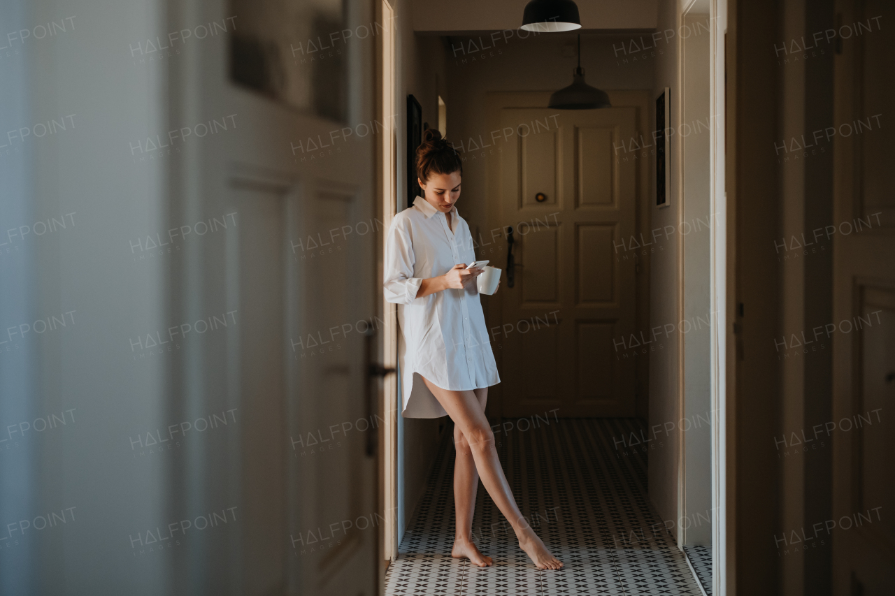 Young woman in pajamas in morning, carrying cup of coffee and returning back to bed. Side view of beautiful single woman at home alone over the weekend.