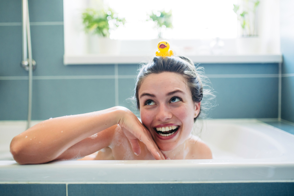 Beautiful young woman in bathtub with rubber duck on head. Enjoying a bubble bath, smiling, having fun.